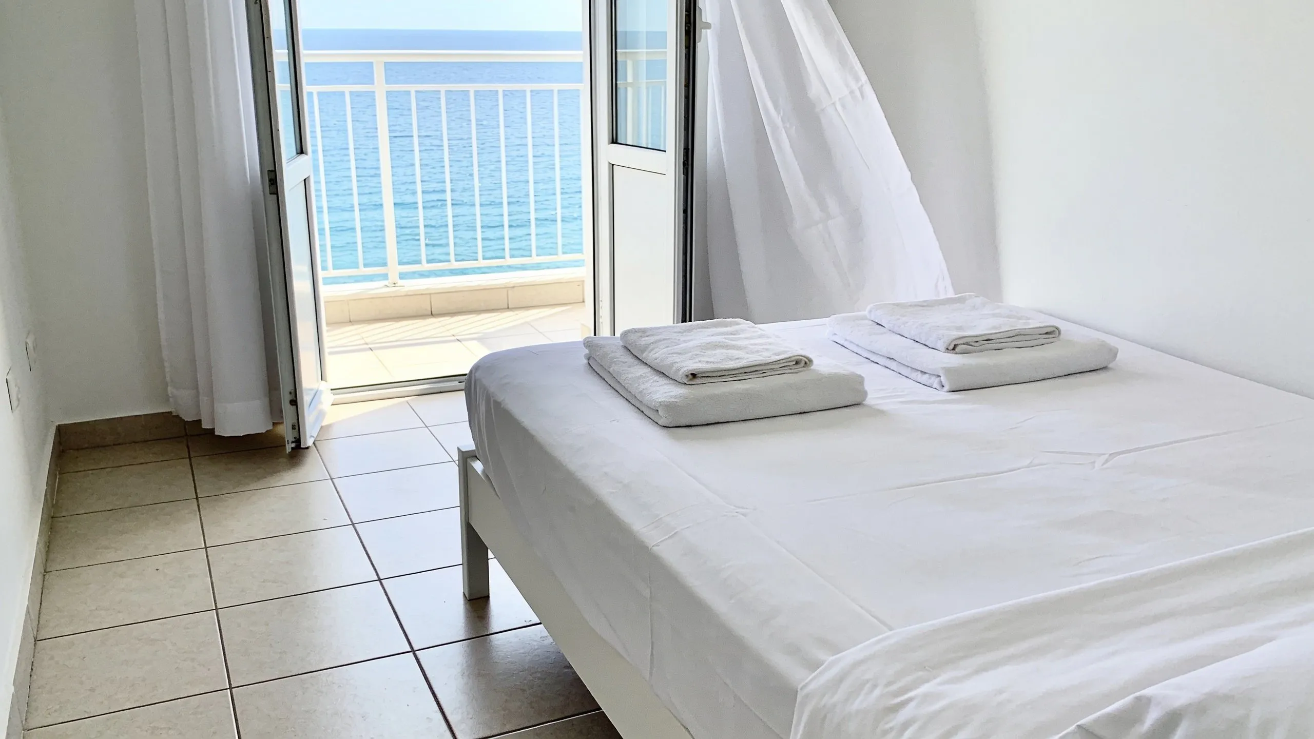 Bedroom of a holiday apartment in Golem, bed made with white sheets and folded towels, clean floor, balcony door open onto the sea